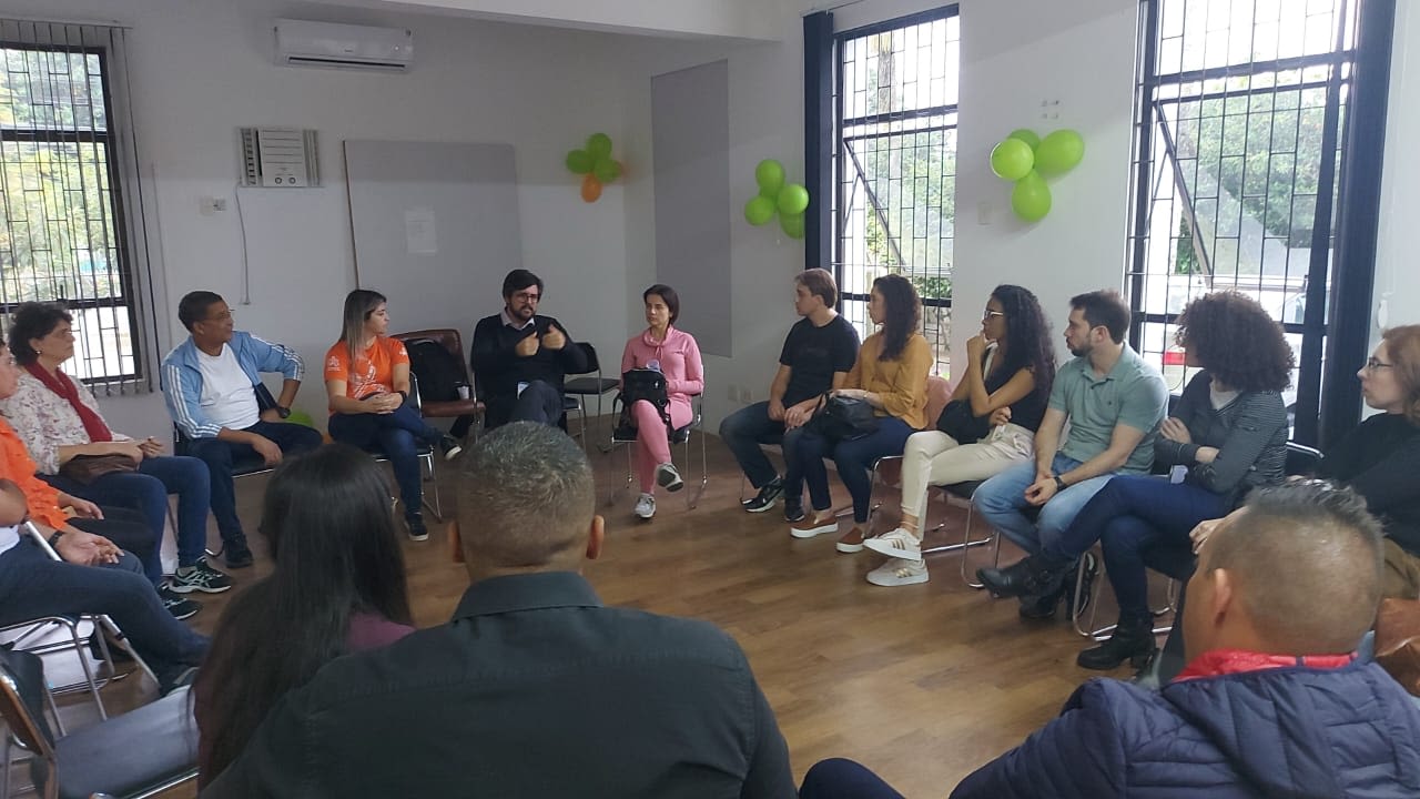 Grupo de 16 pessoas em círculo, sentadas em cadeiras, voltadas para um palestrante que faz gestos com a mão, de frente para o grupo. Estão em uma sala com grandes janelas, balões verdes e laranjas na parede e chão de madeira.